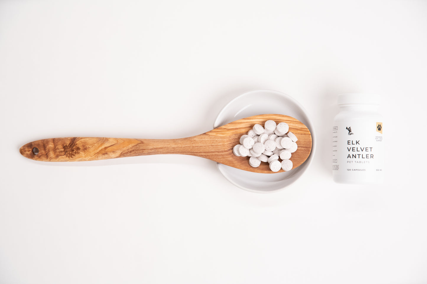 Wooden spoon with white capsules on a white background, next to a white supplement bottle.